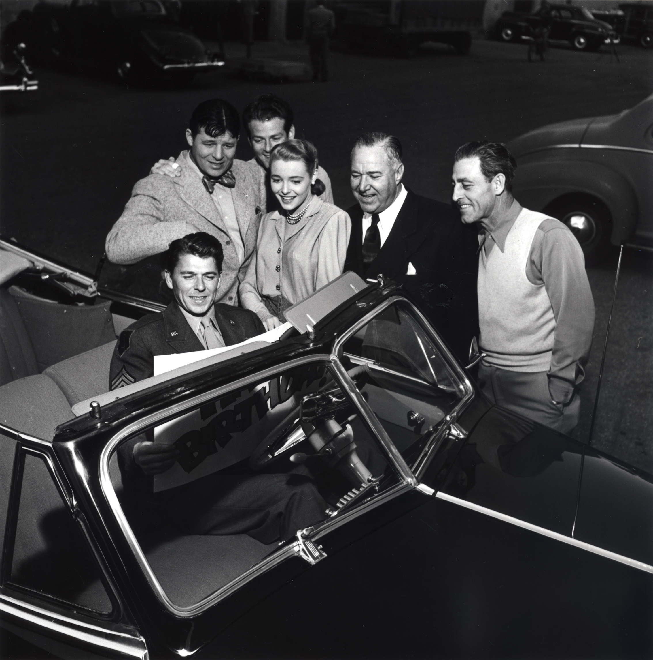 Ronald Reagan, Patricia Neal, Jack Carson, Edward Arnold and 2 unidentified men standing near Reagan who is in a car with a large birthday card