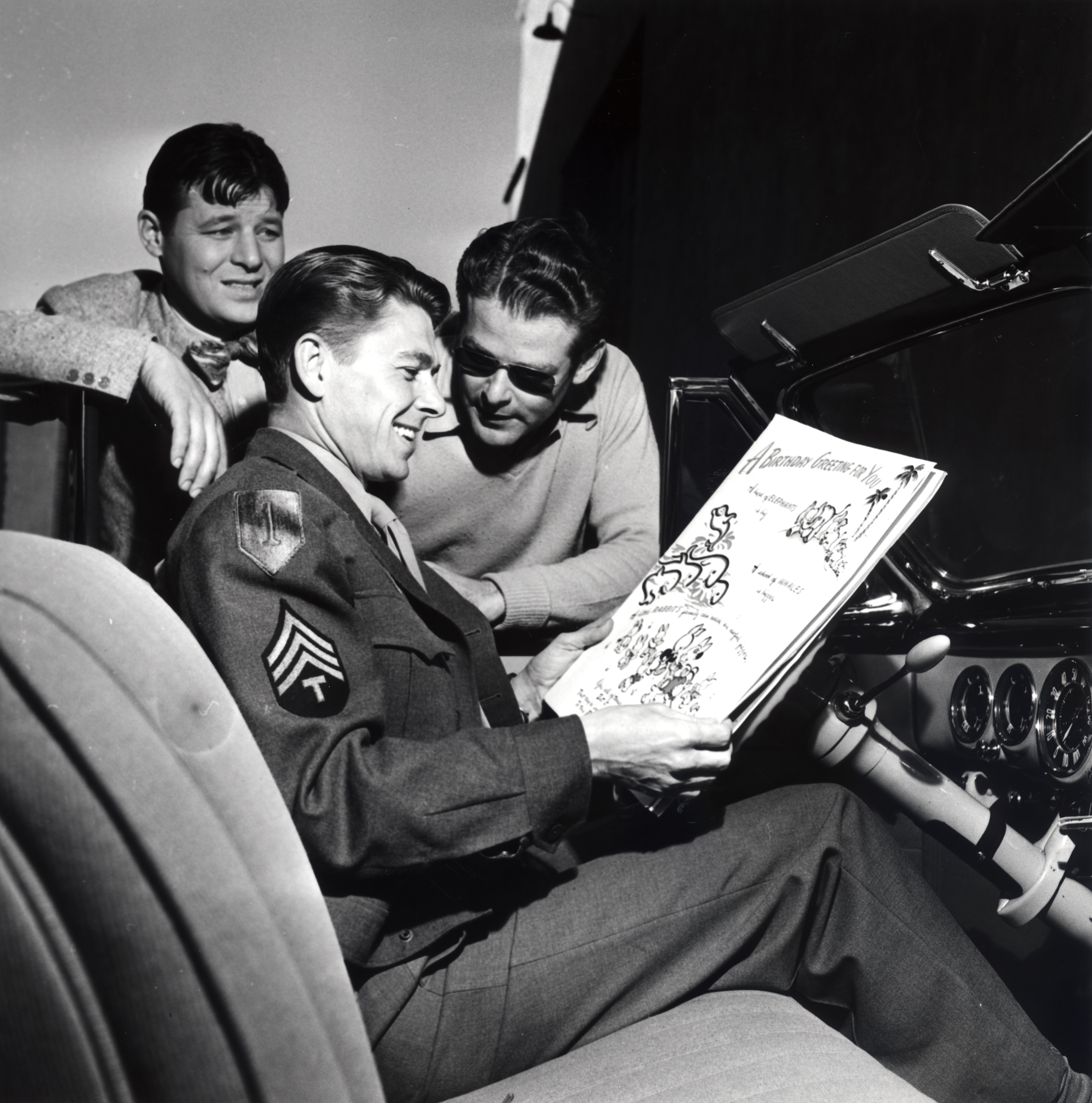 Ronald Reagan, Jack Carson and an unidentified man standing near Reagan who is in a car with a large birthday card