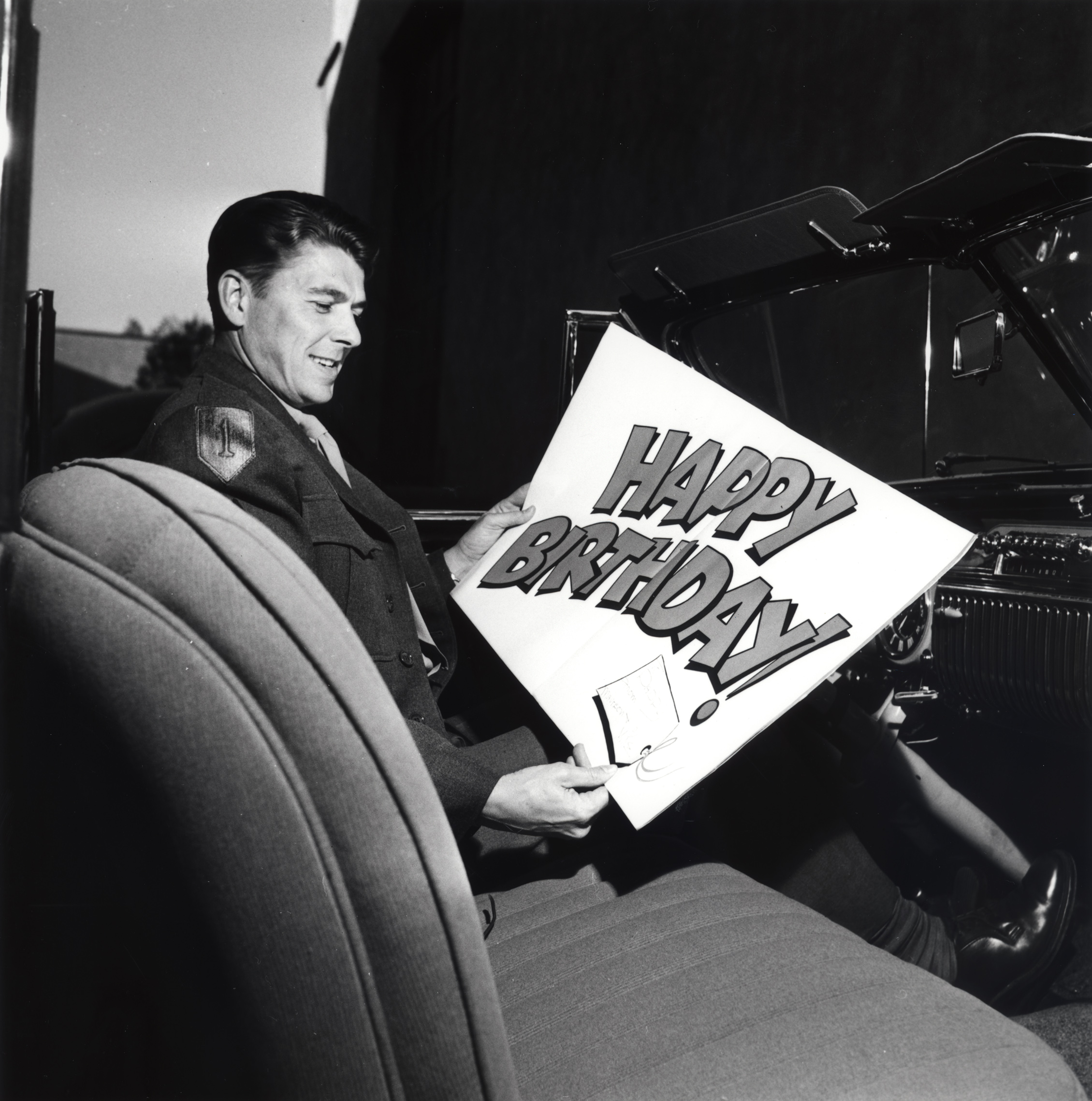 Ronald Reagan sitting a car with a large birthday card