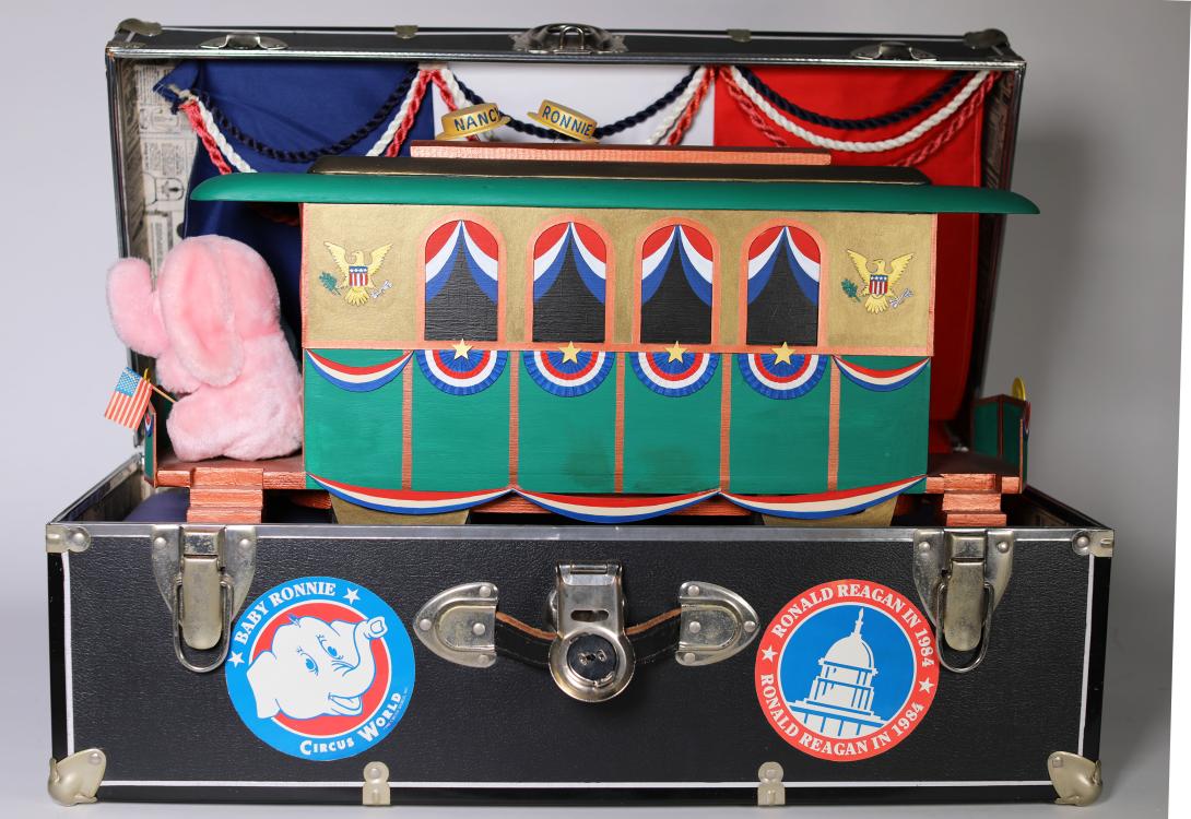 A wooden train car painted green and gold with red, white, and blue detailing. On the front platform are two stuffed elephants. One is pink, and one is blue. The train car sits inside a black trunk covered in 1984 campaign stickers.  