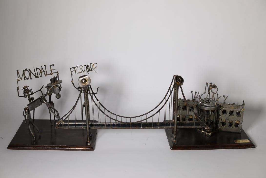 A metal sculpture of a bridge connecting two wooden bases. The base on the left side has three figures representing Mondale and Mrs. Ferraro, along with a donkey attempting to cross the bridge. Above the figures are the words "Mondale and Ferraro." The right base features the White House with the words "No Vacancy" above it. The front corner of the base has a mall brass tone plaque with the text "IF IT DON'T NEED FIXIN'- DON'T FIX IT."