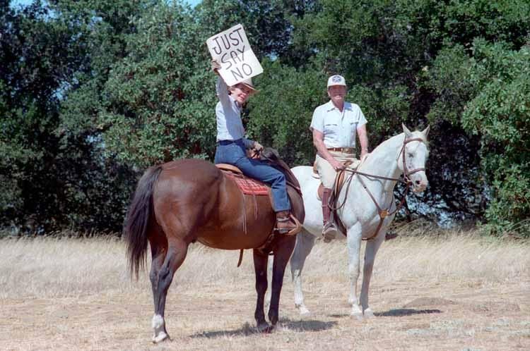 Nancy Reagan holding up “Just Say No” sign for press photographers while horseback riding with President Ronald Reagan at Rancho del Cielo in Santa Barbara, California 09/06/1986  NAID: 75855005