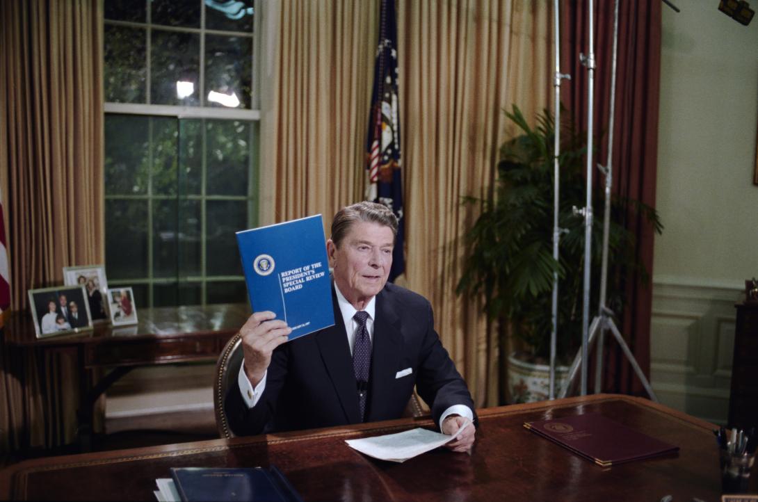 President Ronald Reagan holding up a copy of Report of the President’s Special Review Board during his Address to the Nation on the Iran Arms and Contra Aid Controversy and Administration Goals in the Oval Office 08/12/1987 