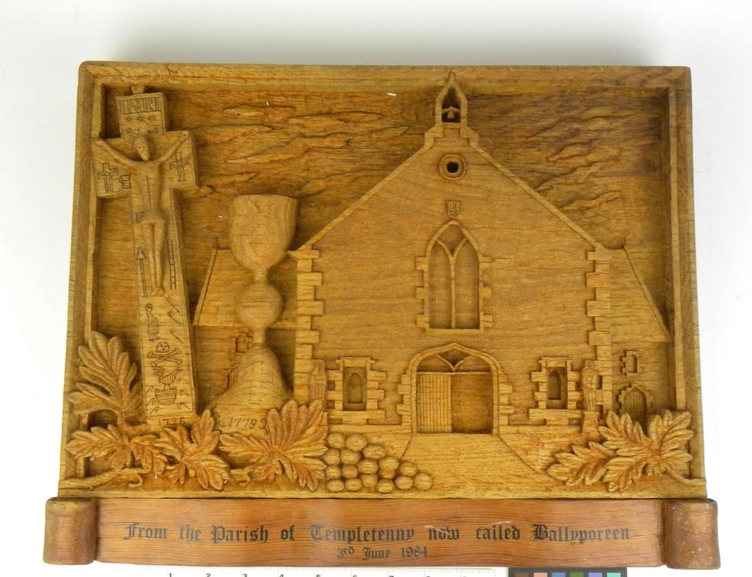 A hand-carved relief made up of multiple religious icons. The left side features the Penal Crucifix, followed by the Templetenny Chalice, and the right side is the 1828 Parish Church of the Assumption in Ballyporeen. Grapevine detailing sits below the carving with text stating, “from the parish of Templetenny now called Ballyporeen 3rd June 1984.”  Gifter: Presented by Denny Walsh on behalf of the People of Ballyporeen Parish. Artifact ID: 1984.01698.01 