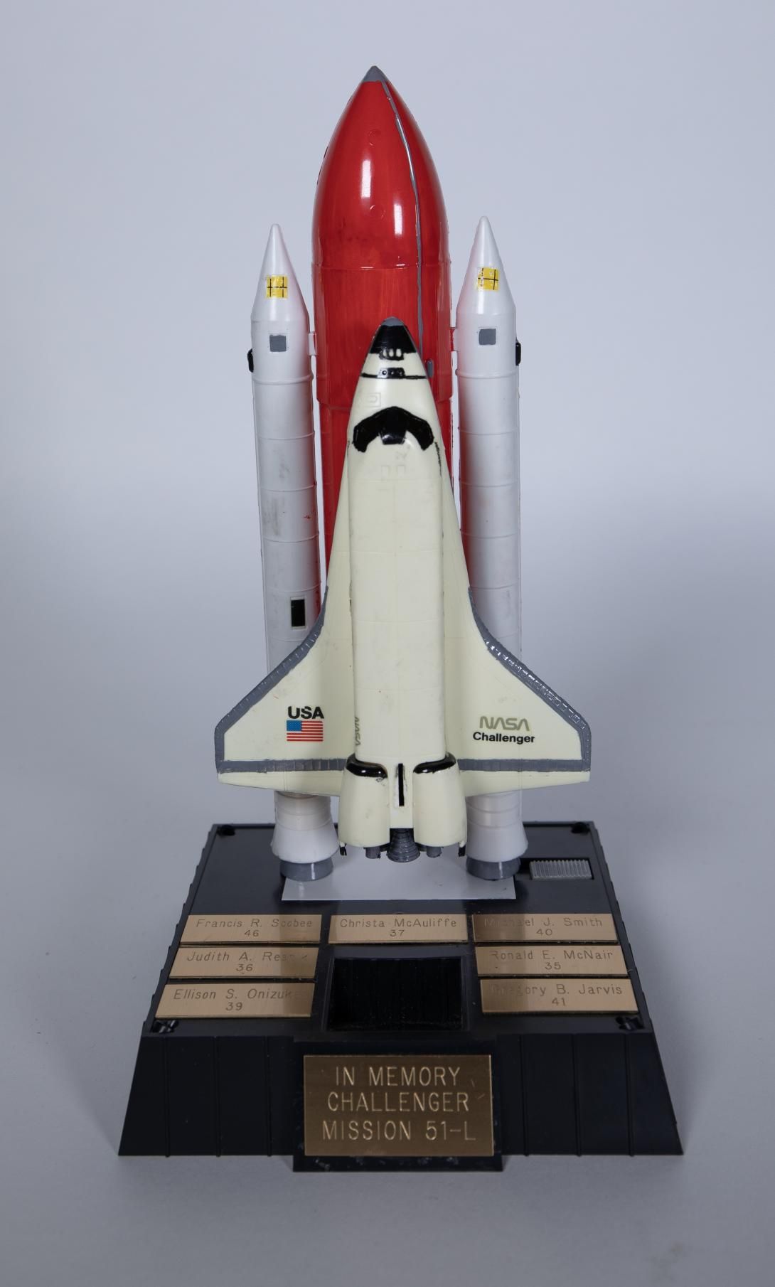  Space Shuttle Challenger Artifact ID: 1986.90806.01a-b Object Name: Model Date Made: 1986 Description: A model of the Space Shuttle Challenger (.01a) with a black display base with seven brass plaques, each bearing the names of the crew members "Francis R. Scobee 46, Christa McAuliffe 37, Michael J. Smith 40, Judith A. Resnik 36, Ronald E. McNair 35, Ellison S. Onizuka 39, Gregory B. Jarvis 41, IN MEMORY CHALLENGER MISSION 51-L." (.01b)