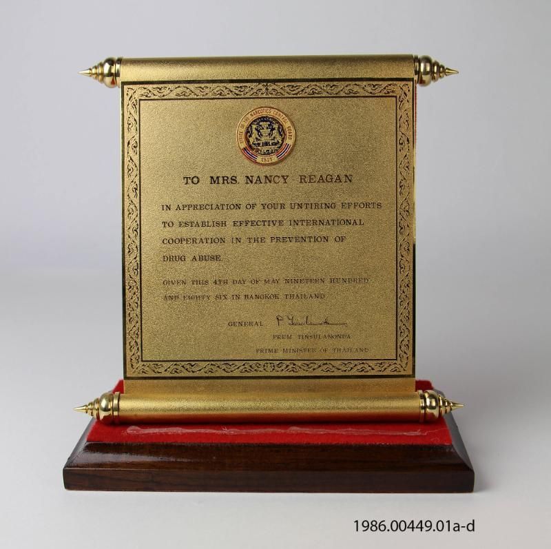 A gold-toned metal scroll (b) mounted on a wooden base (a) The text on the scroll states "TO MRS. NANCY REAGAN IN APPRECIATION OF YOUR UNTIRING EFFORTS TO ESTABLISH EFFECTIVE INTERNATIONAL COOPERATION IN THE PREVENTION OF DRUG ABUSE. GIVEN THIS 4TH DAY OF MAY NINETEEN HUNDRED AND EIGHTY SIX IN BANGKOK, THAILAND GENERAL PREM TINSULANONDA PRIME MINISTER OF THAILAND." At the top of the scroll is an "OFFICE OF THE NARCOTICS CONTROL BOARD" emblem. 