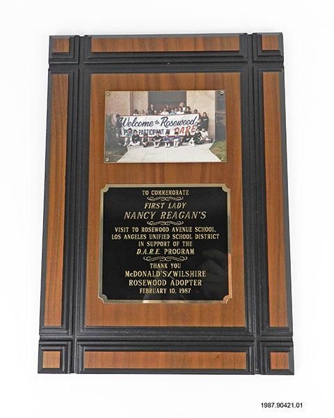 A wooden plaque with a picture of students from Rosewood Avenue School Los Angeles, California, holding a banner stating, "Welcome to Rosewood - A Proud Participant in D.A.R.E." Below the image is a plaque stating, "TO COMMEMORATE FIRST LADY NANCY REAGAN'S VISIT TO ROSEWOOD AVENUE SCHOOL, LOS ANGELES UNIFIED SCHOOL DISTRICT IN SUPPORT OF THE D.A.R.E. PROGRAM - THANK YOU MCDONALD'S/WILSHIRE ROSEWOOD ADOPTER FEBRUARY 10, 1987."
