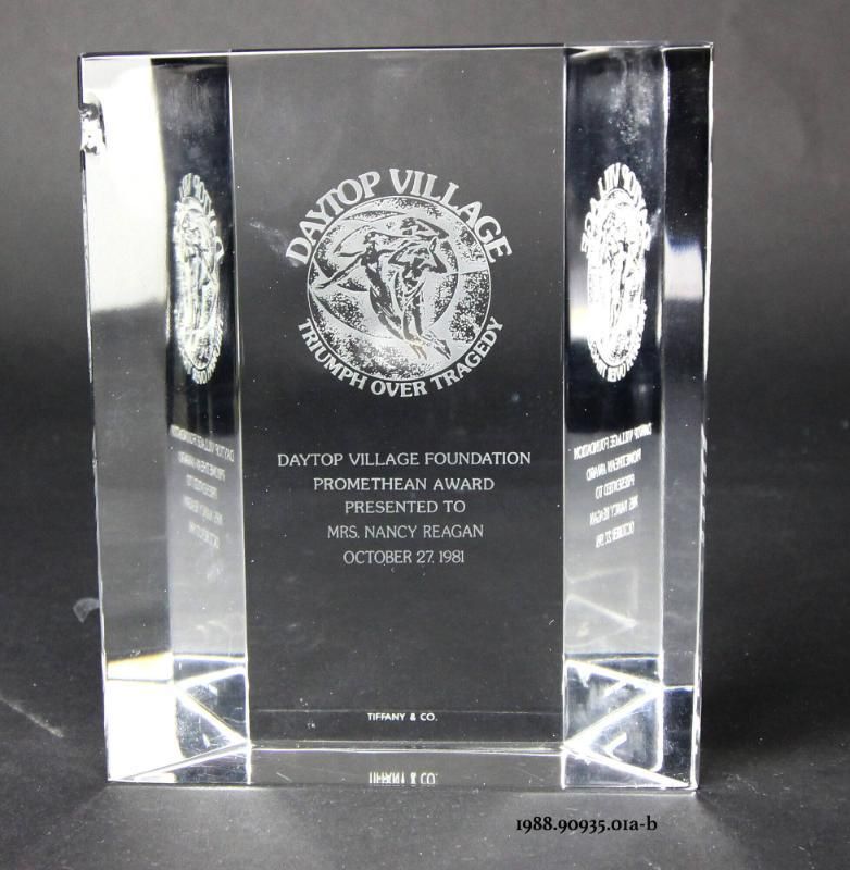 A faceted crystal award reverse etched with a logo of two people flying inside a circle, lettered “DAYTOP VILLAGE TRIUMPH OVER TRAGEDY” around the edges, and “DAYTOP VILLAGE FOUNDATION PROMETHEAN AWARD PRESENTED TO MRS. NANCY REAGAN OCTOBER 27, 1981” in the center and “TIFFANCY & CO.” at the bottom. Presented to First Lady Nancy Reagan when she was in New York City, New York in 1981 for the Daytop Village presentation on October 26 and the Daytop Dinner on October 27. 