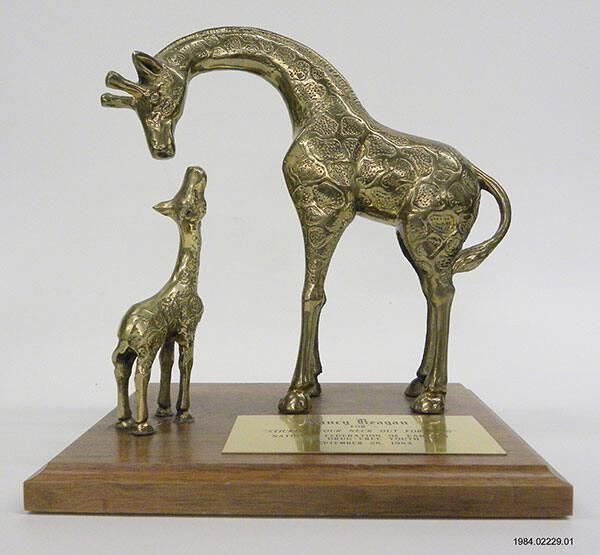 This is a sculpture of two brass giraffes, one large and the other small. The larger giraffe is craning its neck down toward the smaller giraffe, which is looking up toward it. The giraffes are mounted on a wooden base with a brass plaque stating "NANCY REAGAN 'FOR STICKING YOUR NECK OUT FOR KIDS', NATIONAL FEDERATION OF PARENTS FOR DRUG-FREE YOUTH, SEPTEMBER 28, 1984." Nancy Reagan attended the third annual National Federation of Parents NFP luncheon on 09/28/1984.